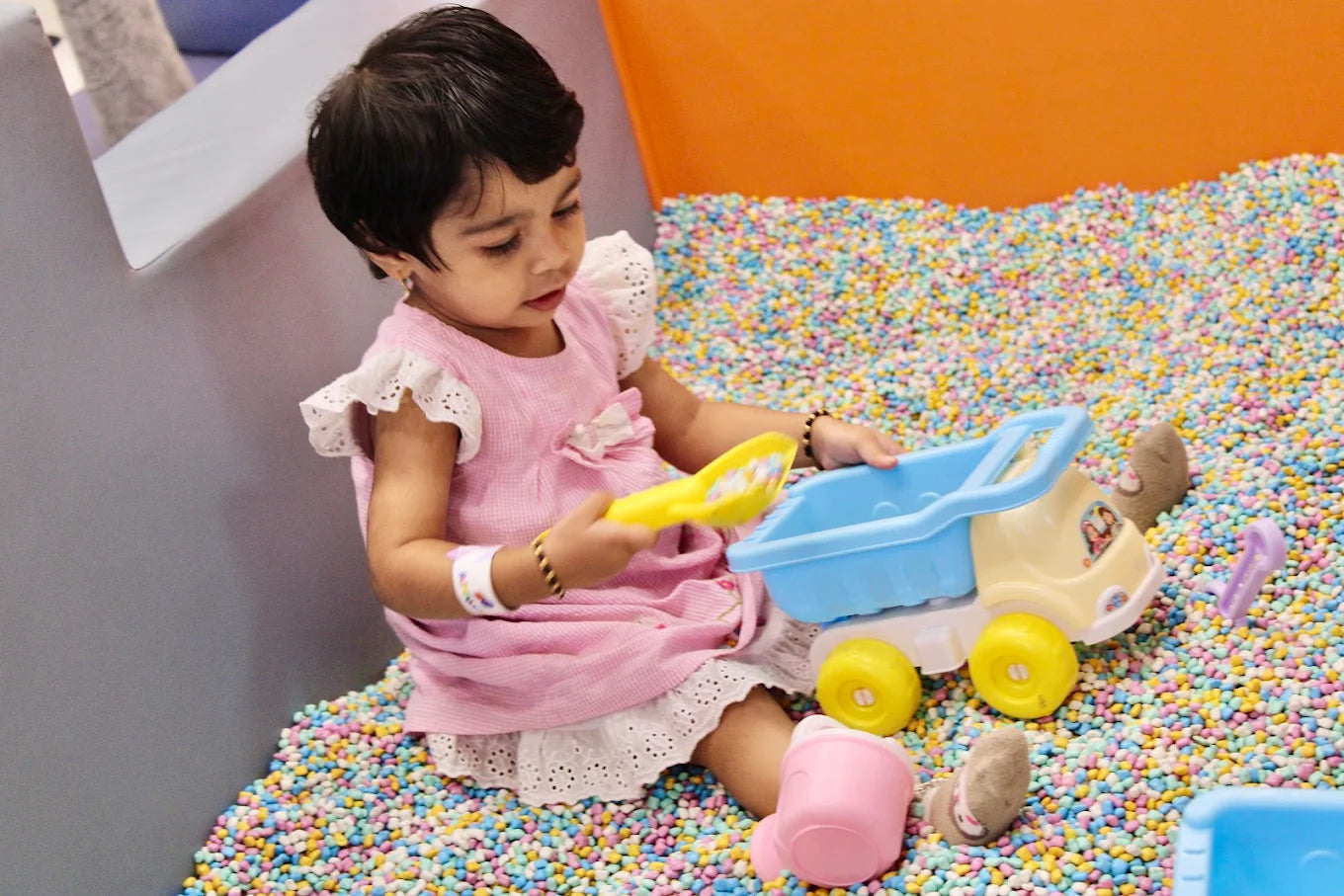 A young child plays with a toy truck and shovel in a ball pit filled with colorful balls.