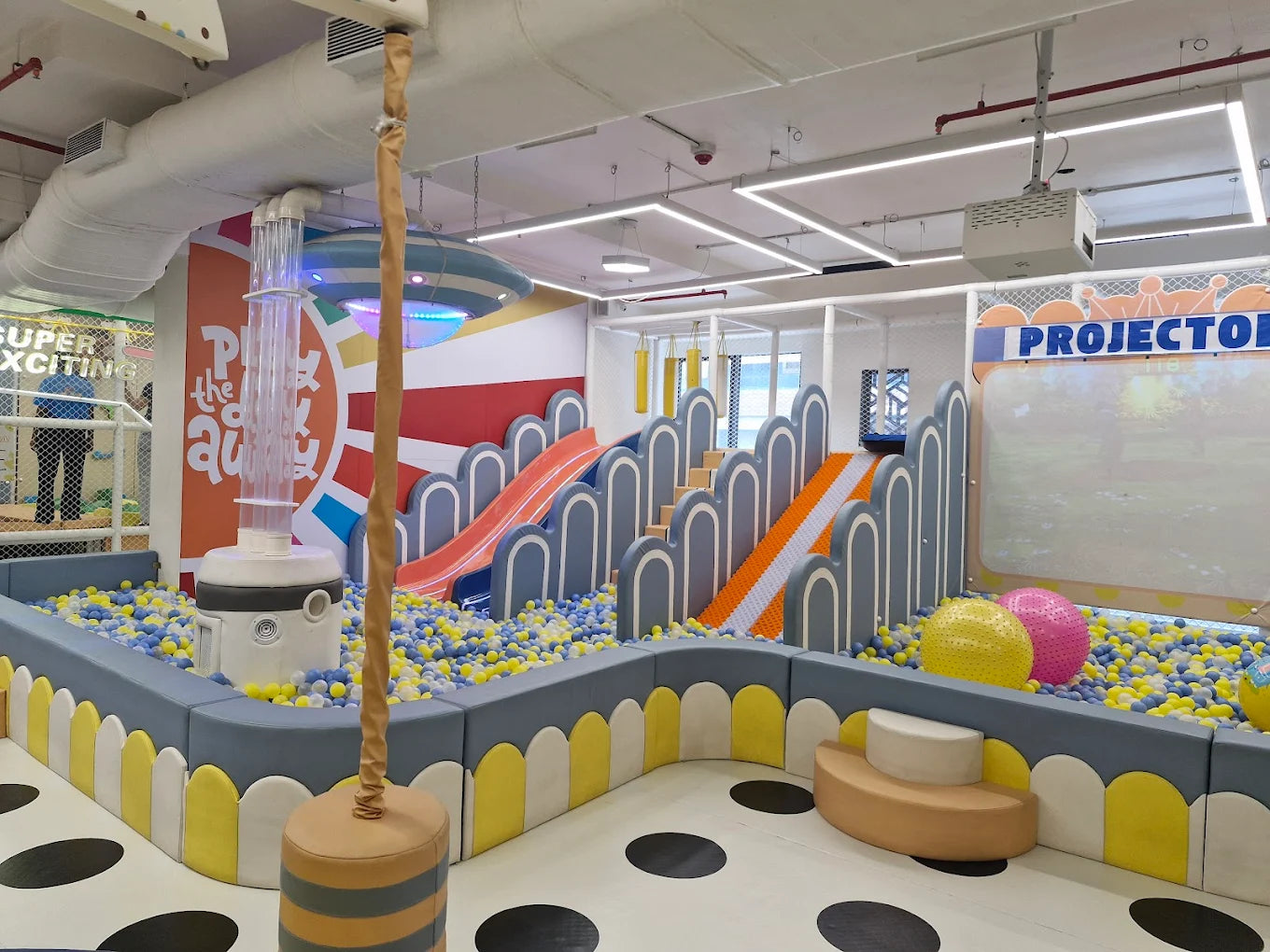 A colorful indoor playground with ball pits, slides, and a projector screen for children.