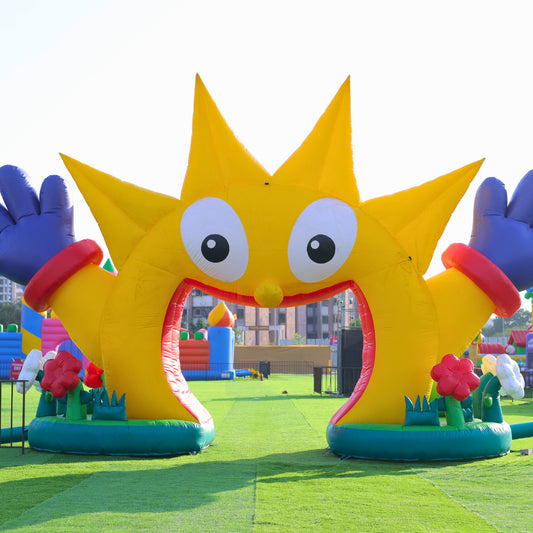 A large, yellow inflatable character with big eyes stands at the entrance to a children's play area. The character has blue hands and is decorated with red flowers.