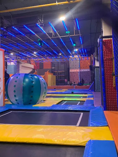 An indoor trampoline park with colorful padded areas and blue overhead lights, where children are playing.