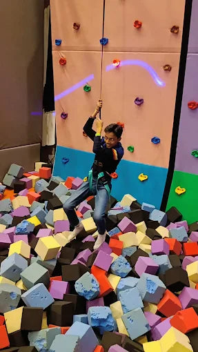 A young person rappels down a climbing wall into a foam pit.