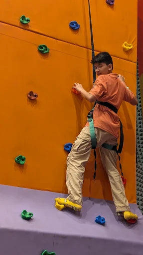 A young person climbs an orange rock-climbing wall, wearing a harness and tan pants.