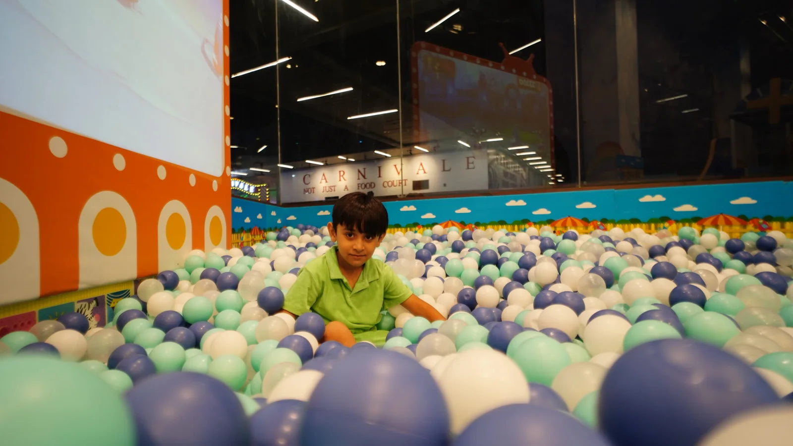 A young boy sits in a ball pit filled with blue, white, and green balls. The background shows a "Carnivale" sign and a colorful wall.
