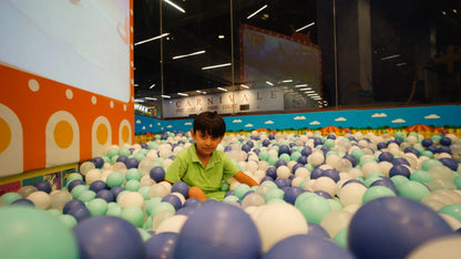 A young boy sits in a ball pit filled with blue, white, and green balls. The background shows a "Carnivale" sign and a colorful wall.