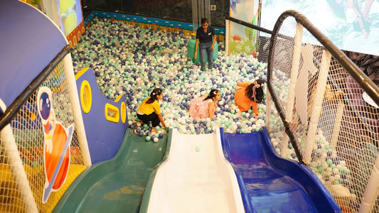 Children play in a ball pit with slides, supervised by an adult.