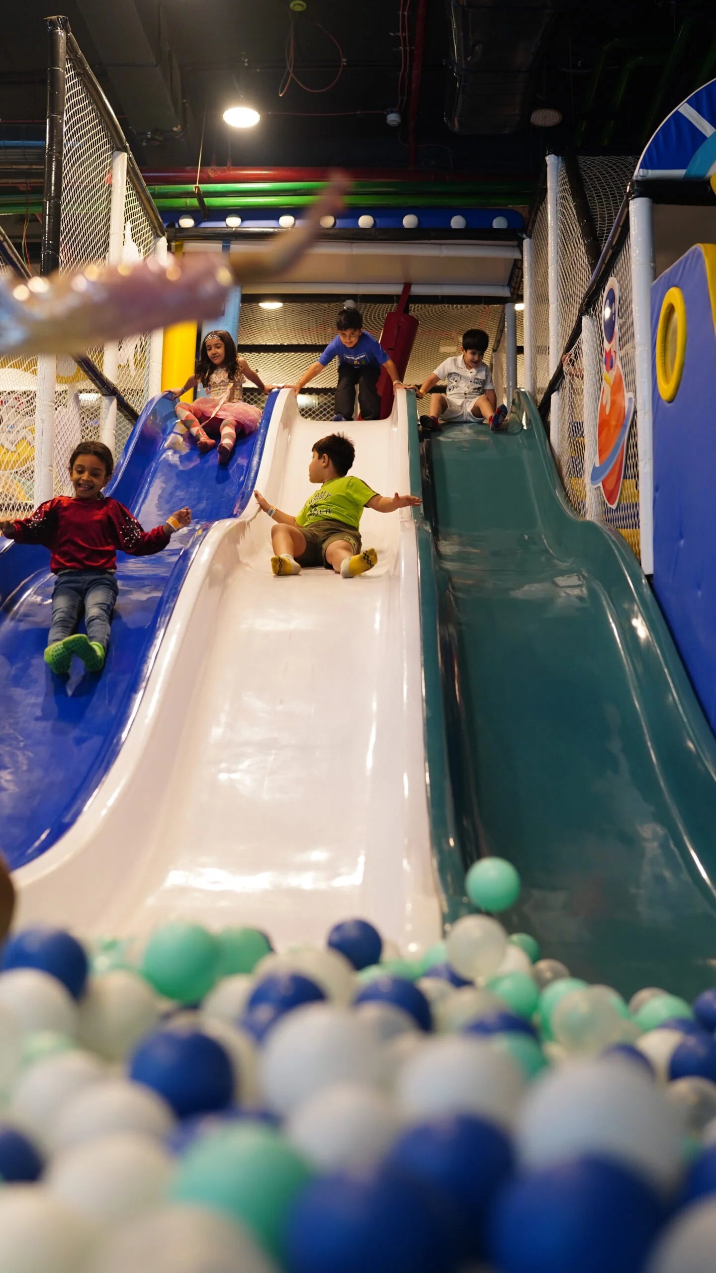 Children slide down a colorful indoor playground slide into a ball pit.
