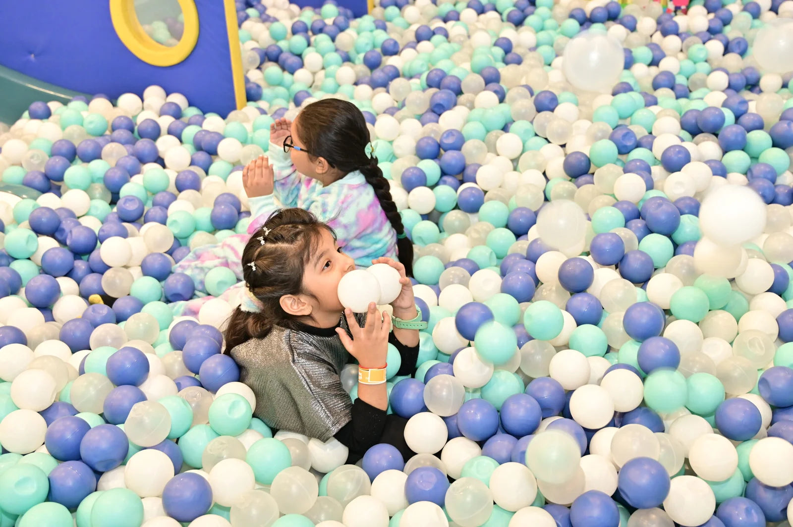 Two young girls play in a ball pit filled with blue, white, and teal balls.