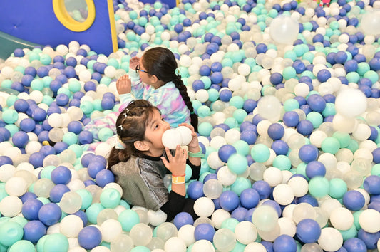 Two young girls play in a ball pit filled with blue, white, and teal balls.