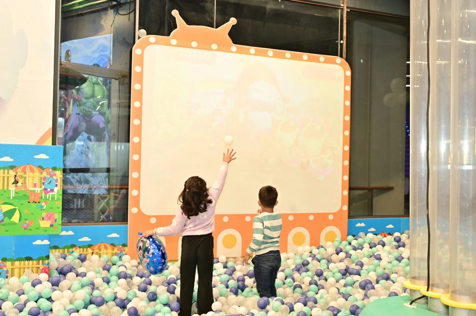 Two young children play in a ball pit in front of a large screen, with one child throwing a ball.