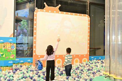 Two young children play in a ball pit in front of a large screen, with one child throwing a ball.