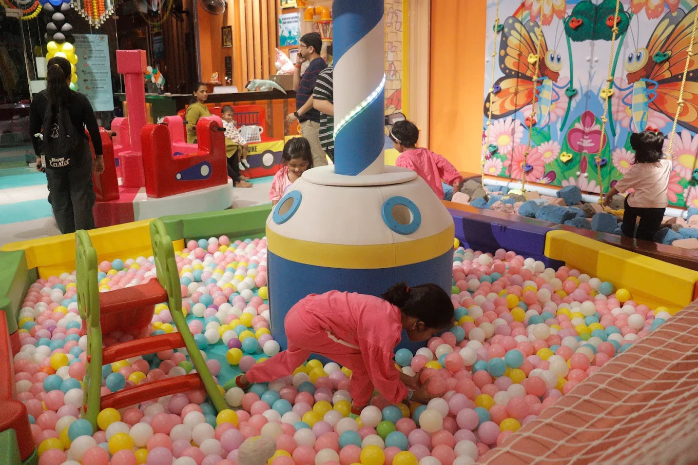Children play in a ball pit with a large, colorful structure in the center. A woman watches from the side.