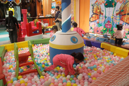 Children play in a ball pit with a large, colorful structure in the center. A woman watches from the side.