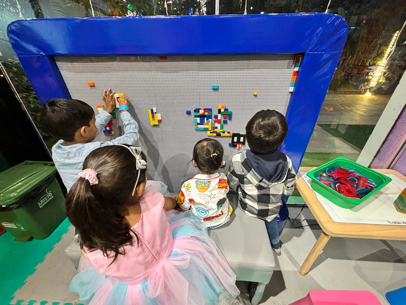 Children build with colorful LEGOs on a large wall in a playful indoor setting.