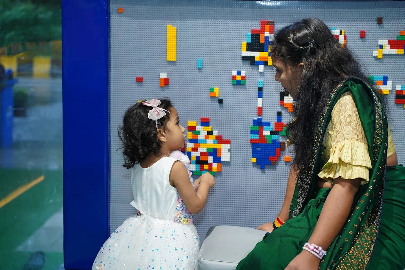 A young girl and a woman look at a wall covered in colorful Lego creations.