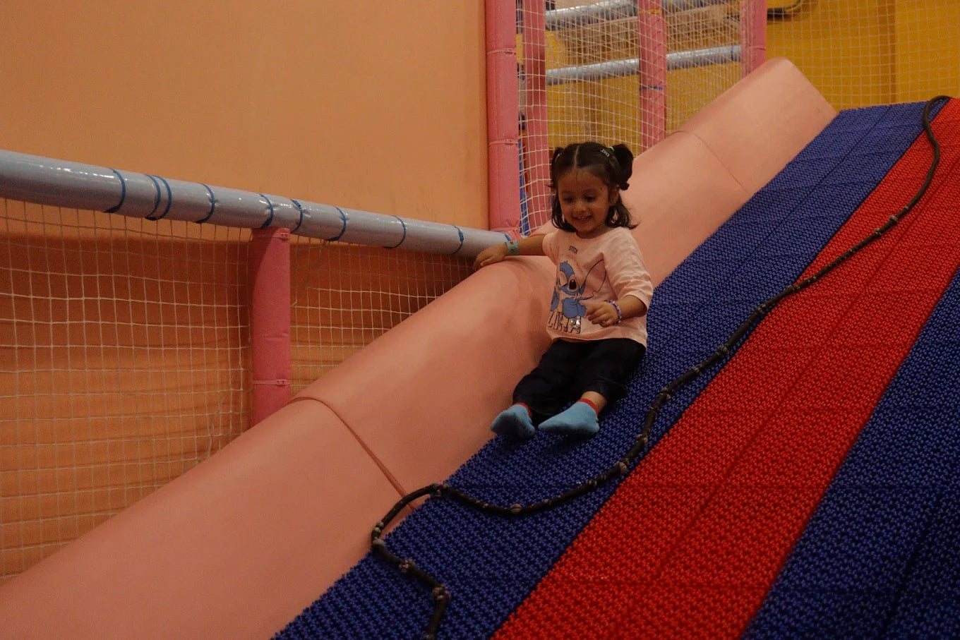 A young girl slides down a colorful indoor playground slide. She is smiling and wearing a pink shirt with a cartoon character.