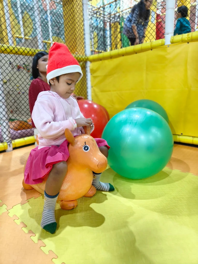 A young child in a Santa hat rides an orange toy horse in a colorful indoor play area.