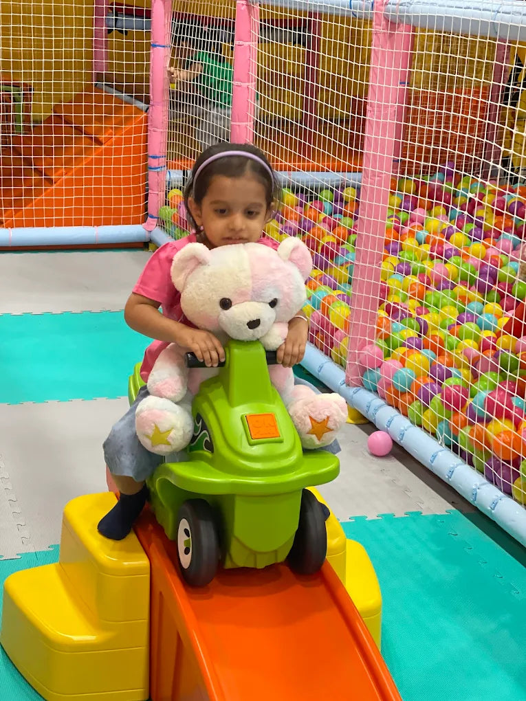A young girl rides a green toy car with a teddy bear in a colorful indoor playground.