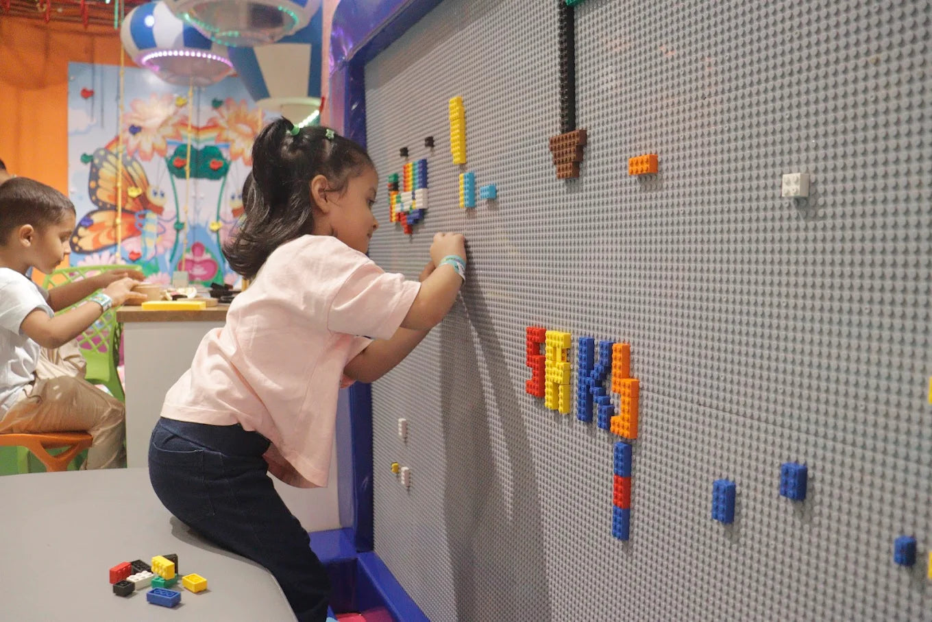 A young girl and boy play with colorful building blocks on a large gray pegboard wall in a brightly decorated room.