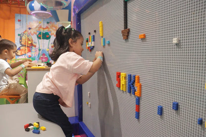 A young girl and boy play with colorful building blocks on a large gray pegboard wall in a brightly decorated room.