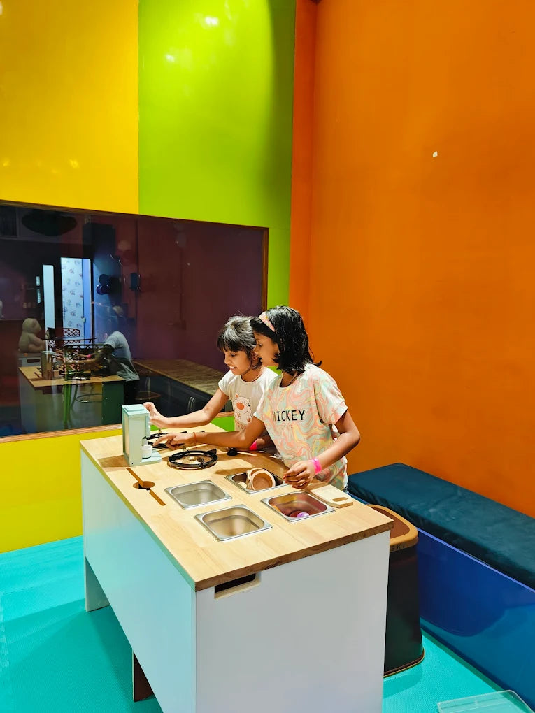 Two young girls play at a toy kitchen in a brightly colored room.