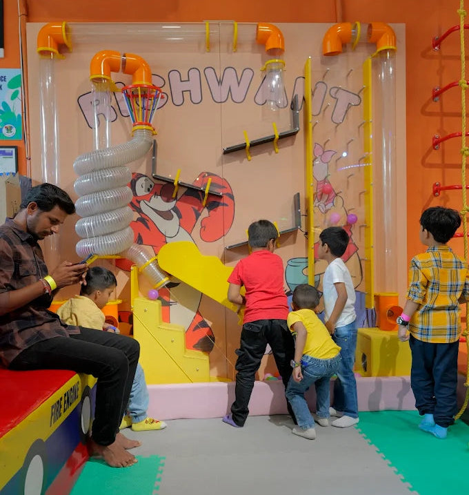 Children play with a ball-dropping tube system in an indoor playground. A man watches them.