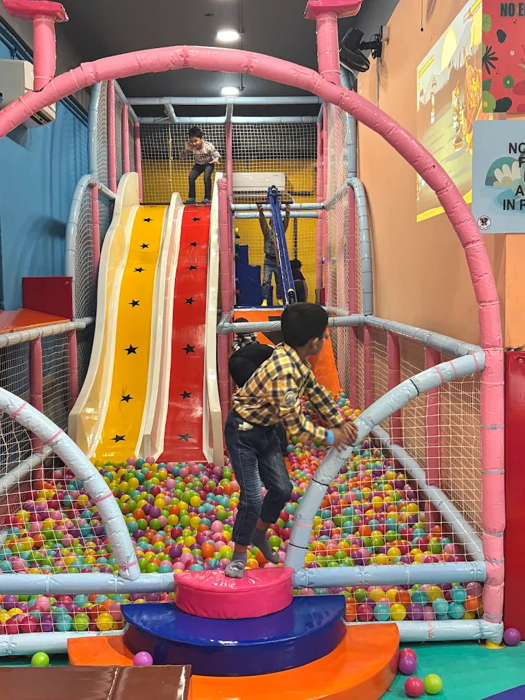 Children play in a colorful ball pit with slides at an indoor playground.