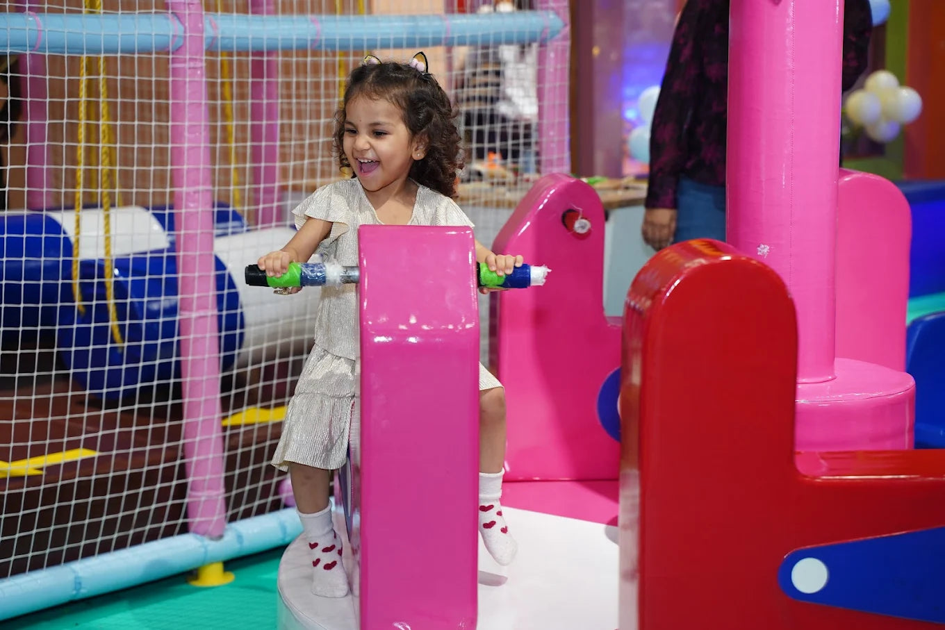 A young girl laughs while playing on a pink ride in an indoor playground.