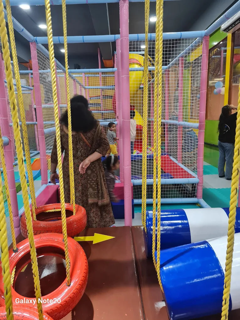 Children play in a colorful indoor playground with ropes and soft obstacles. A woman watches them from the side.