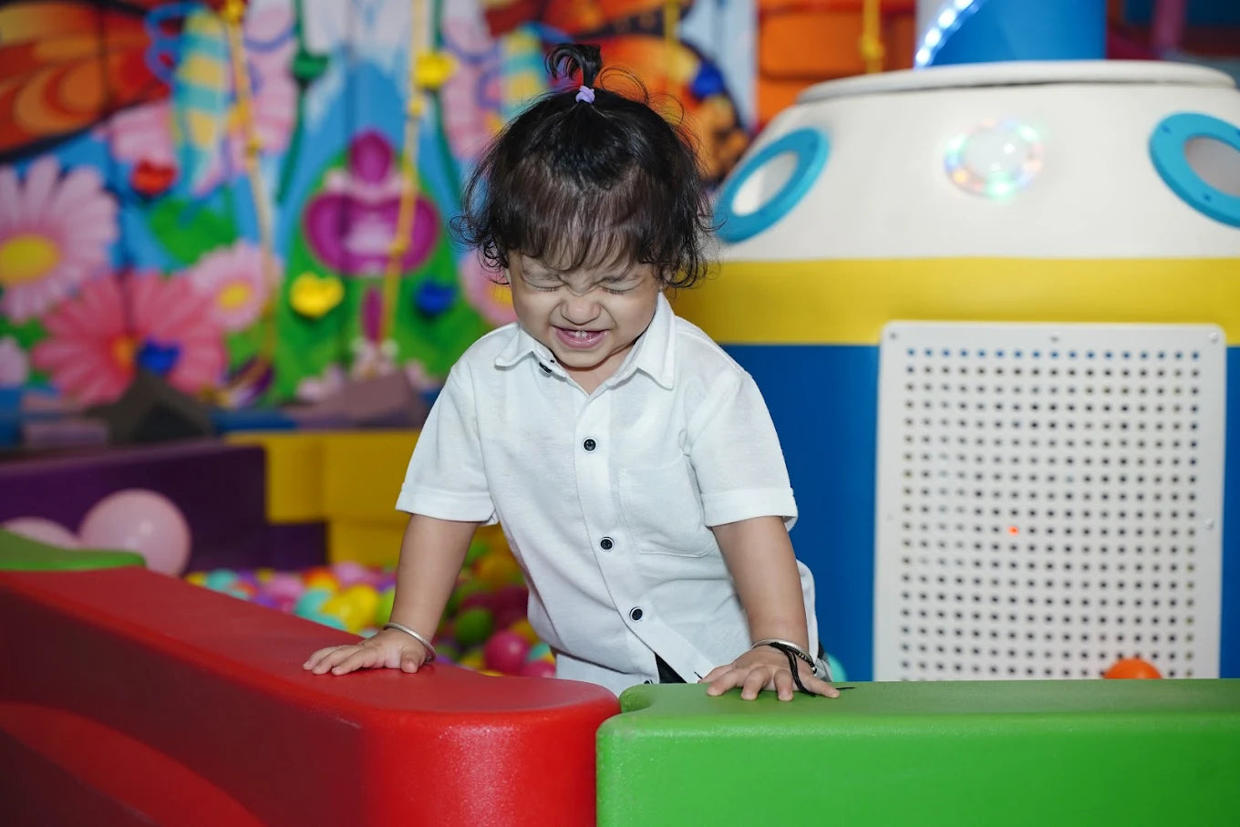 A young child with a playful expression leans on colorful play structures in an indoor playground.