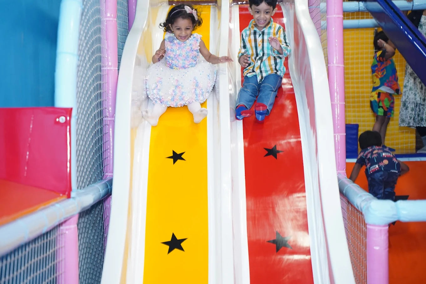 Two young children slide down a colorful indoor playground slide, one on a yellow path and the other on a red path.