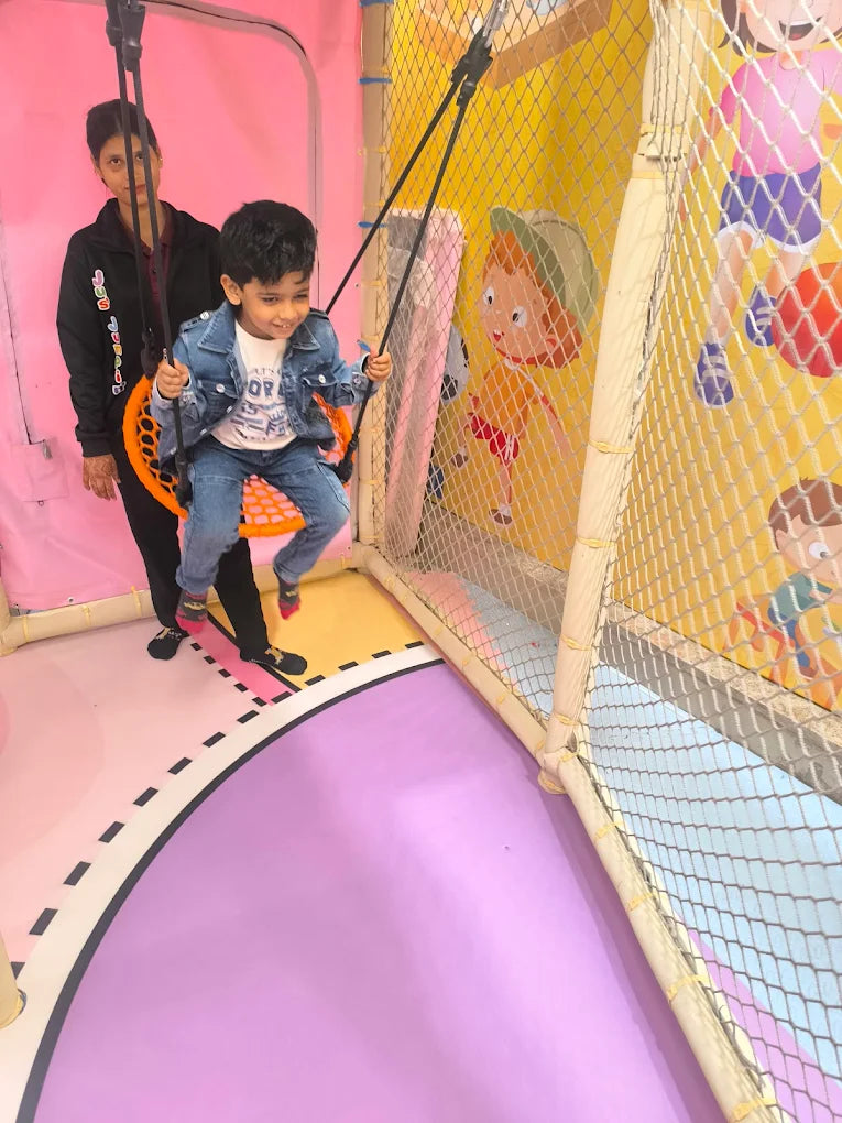 A young boy on a swing is pushed by an adult in a colorful indoor play area.