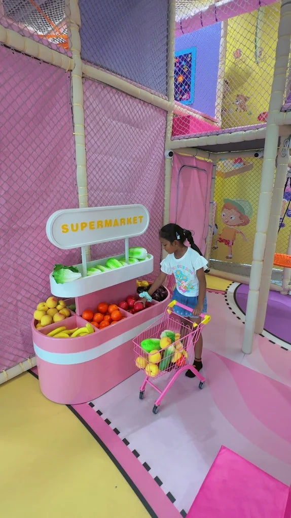 A young girl shops at a toy supermarket, filling her pink cart with plastic fruits and vegetables.