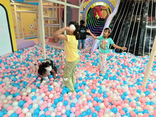 Children play in a ball pit with pink, blue, and white balls, with a slide in the background.
