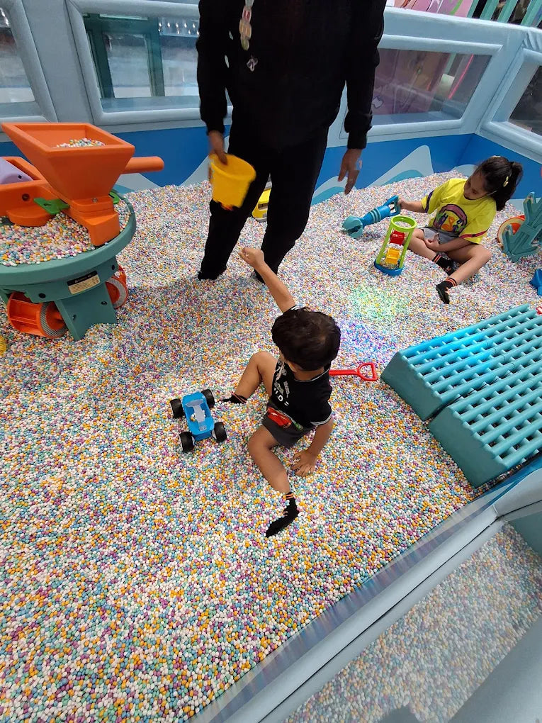 A young child plays with a blue toy car in a ball pit while an adult and another child are visible in the background.