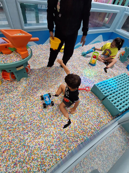 A young child plays with a blue toy car in a ball pit while an adult and another child are visible in the background.