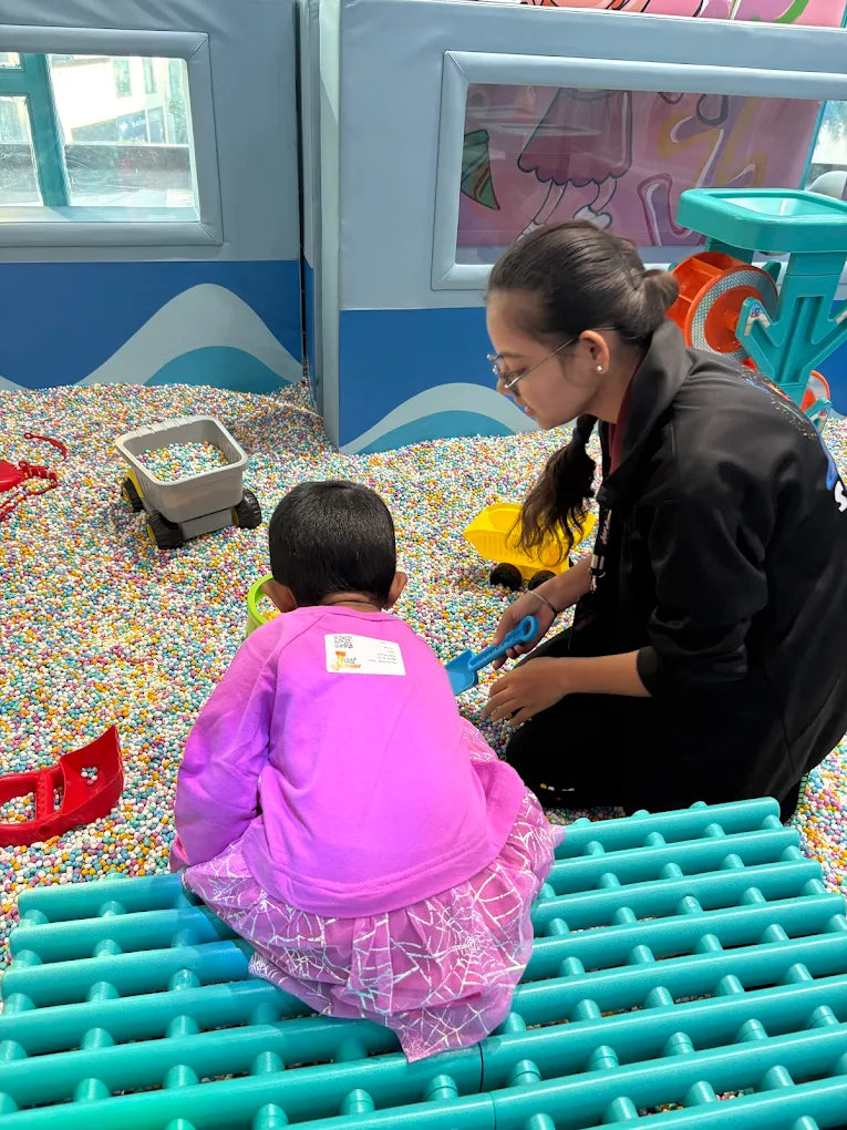 A young child plays in a ball pit with an adult, surrounded by colorful balls and toys.