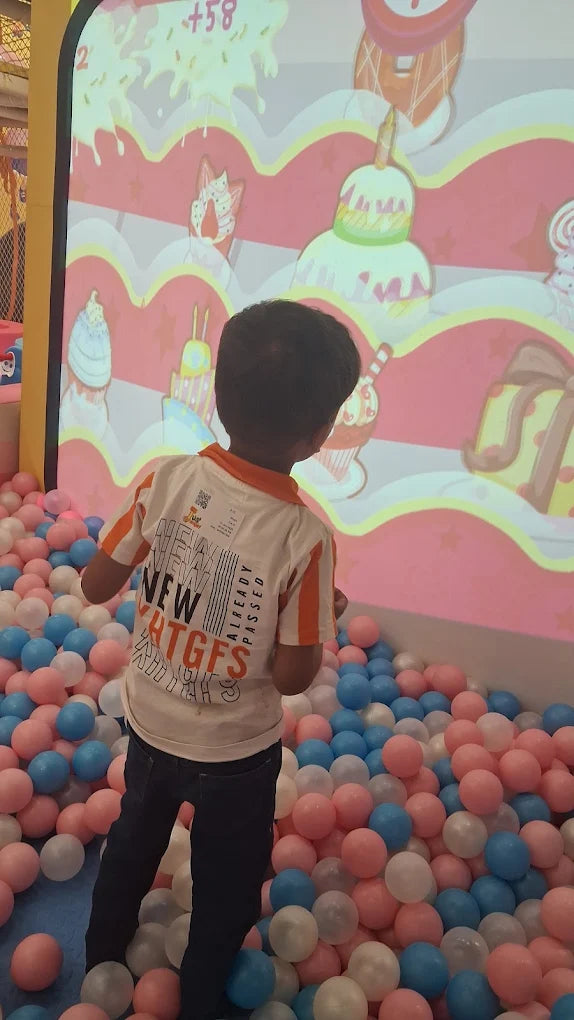 A young child plays in a ball pit in front of a projected game with cakes.