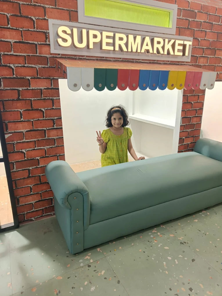 A young girl stands in a play supermarket, making a peace sign. A teal bench sits in front of the play store.