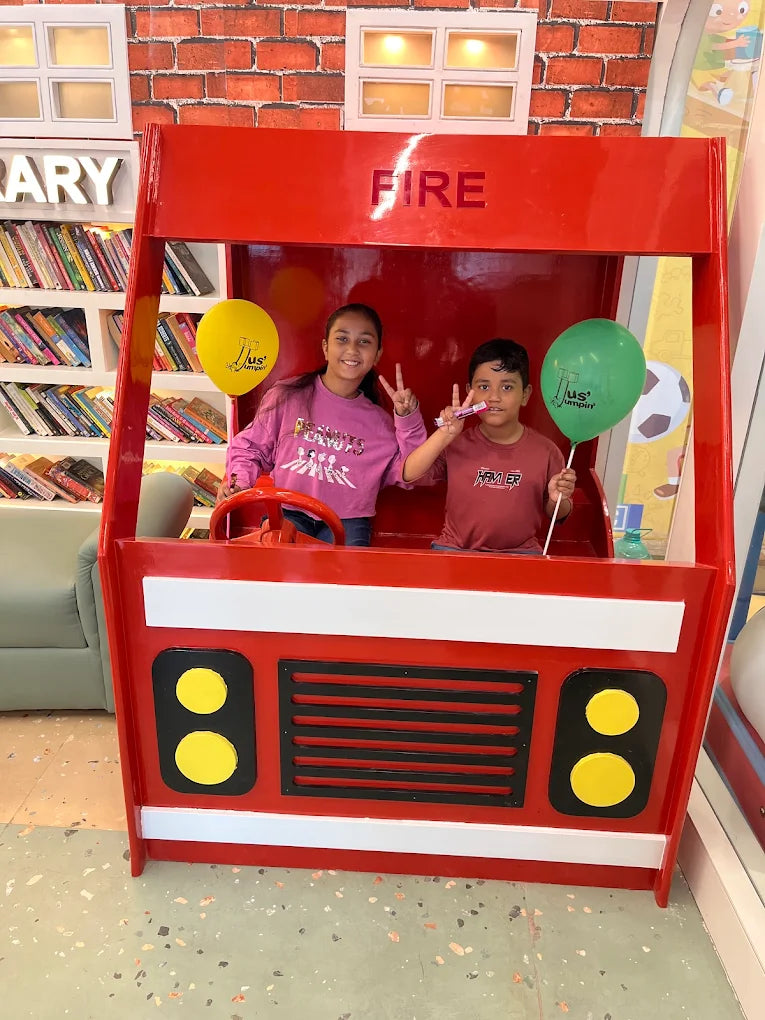 Two children pose in a red fire truck playhouse with balloons.