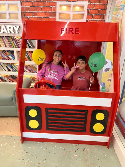 Two children pose in a red fire truck playhouse with balloons.