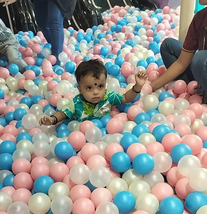 A young child sits in a ball pit filled with pink, blue, and white balls. The child is wearing a green and white patterned shirt.