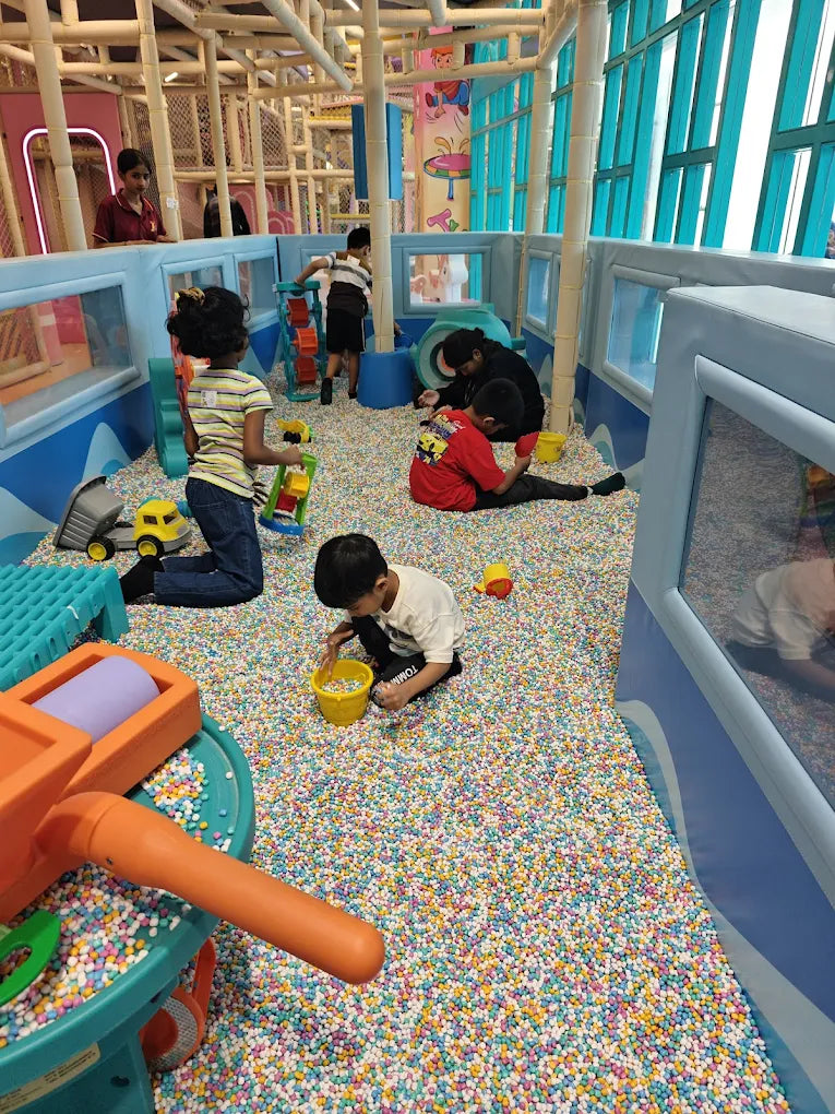 Children play in a ball pit at an indoor playground. A young boy scoops balls into a yellow bucket.