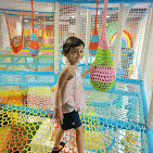 A young child stands in a colorful indoor playground with nets and soft play structures.