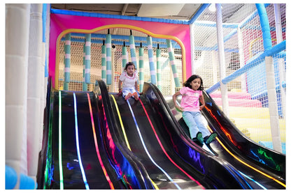 Two young girls slide down a colorful, illuminated playground slide.