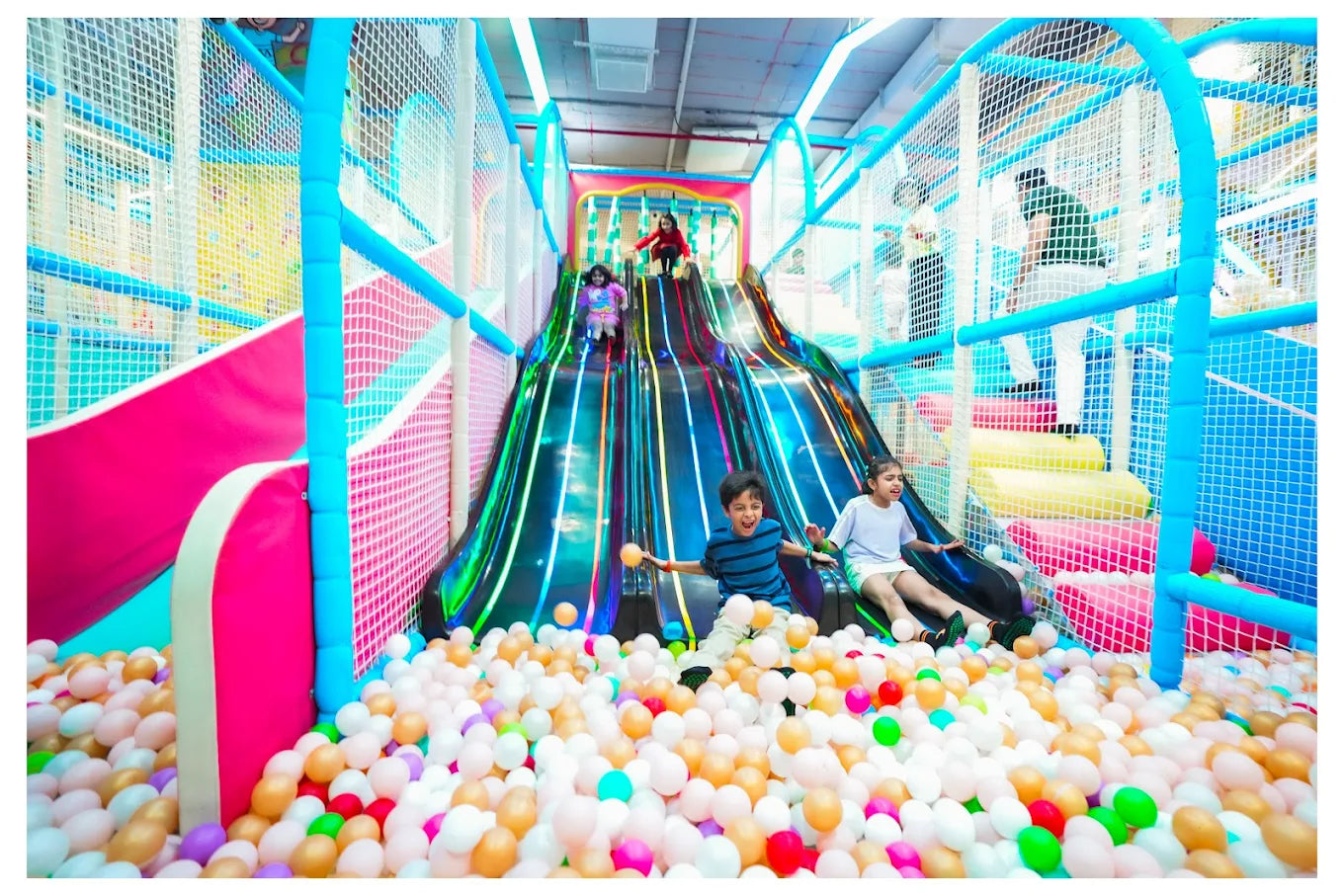 Children slide down a colorful indoor playground slide into a ball pit.