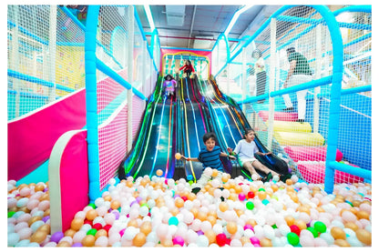Children slide down a colorful indoor playground slide into a ball pit.