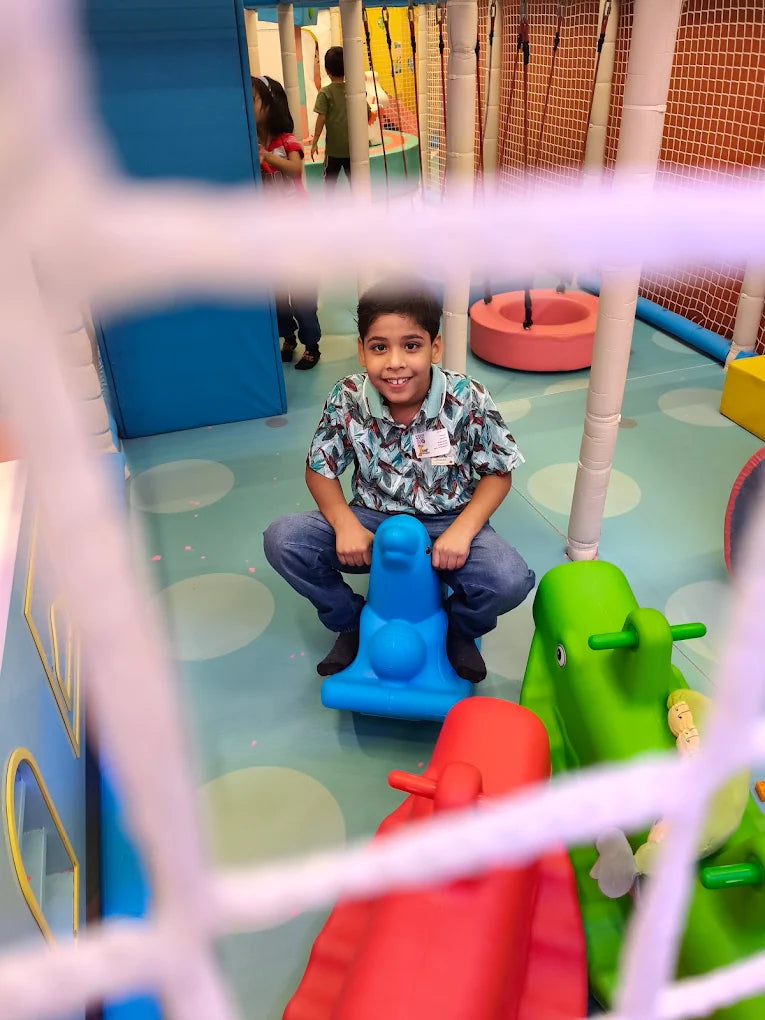 A young boy smiles while sitting on a blue rocking toy in a colorful indoor playground. Other children and play structures are visible in the background.