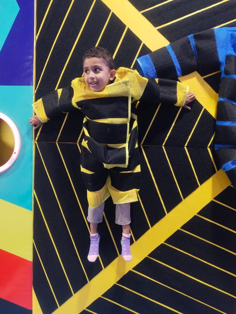 A young child in a bee costume is suspended on a climbing wall with black and yellow stripes.