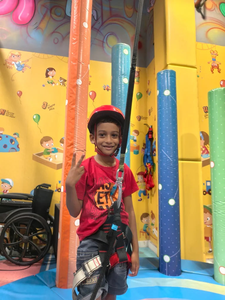 A young boy in a red helmet and harness smiles at the camera, ready to climb an indoor climbing wall.
