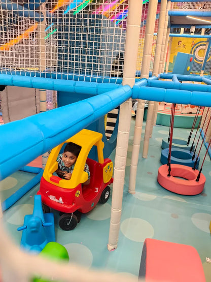 A young boy smiles from inside a toy car at an indoor playground. The colorful play area features soft flooring and climbing structures.